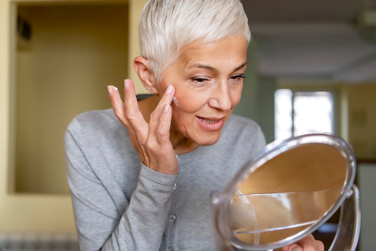 Older woman with short gray hair applies cream to her face while looking into a handheld mirror indoors, focusing on her summer skincare routine.