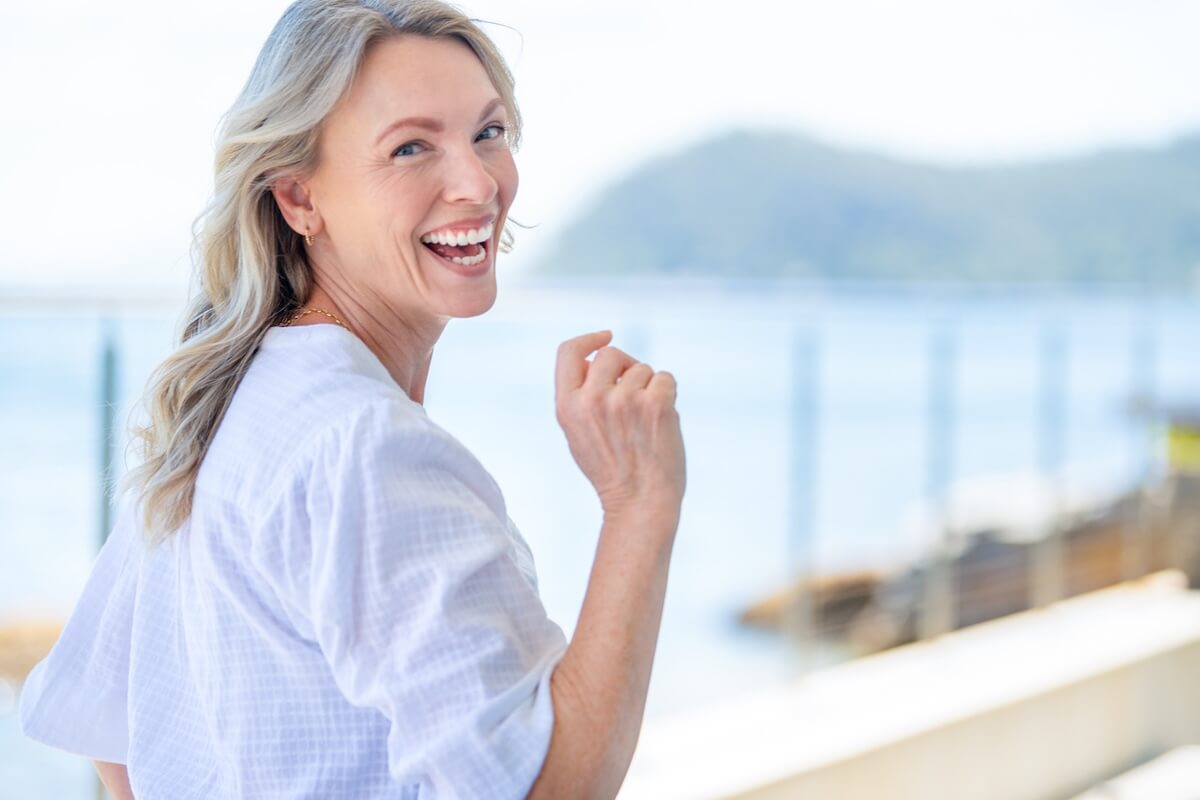 A woman with blonde hair in a white shirt smiles while looking back outdoors near a waterfront, her glowing complexion hinting at a dedicated summer skincare routine amid blurred mountains in the background.