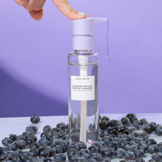The 5 Best-Smelling Skincare Products I Can't Live Without - 5 A hand presses the pump of a clear bottle labeled "Blueberry Bounce Gentle Cleanser" by Glow Recipe—one of the best-smelling skincare products—surrounded by scattered blueberries on a light surface with a purple background.