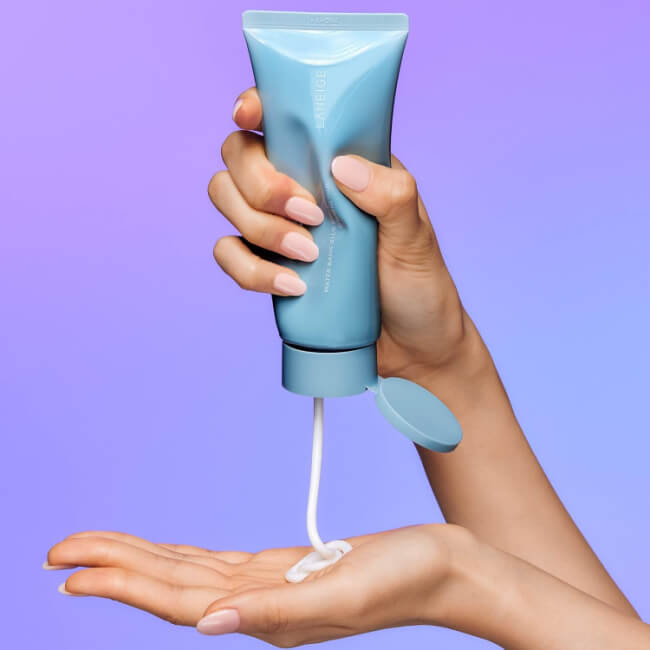 The 5 Best-Smelling Skincare Products I Can't Live Without - 2 A hand squeezes white lotion from a blue tube onto the palm of another hand against a purple background, showcasing the best-smelling skincare in action.