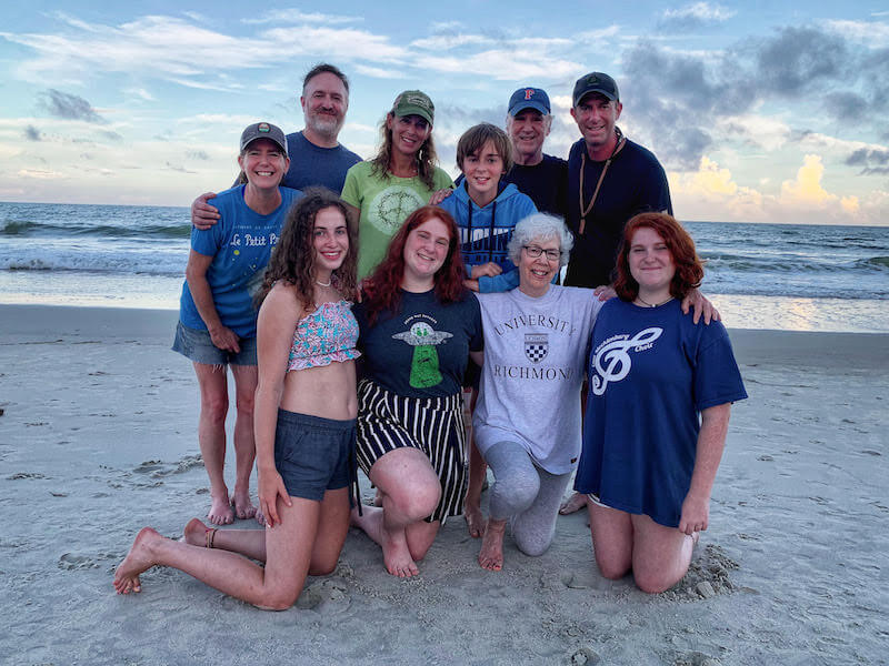 A group of ten people, including adults and teenagers, pose together on a sandy beach with the ocean and a cloudy sky in the background.