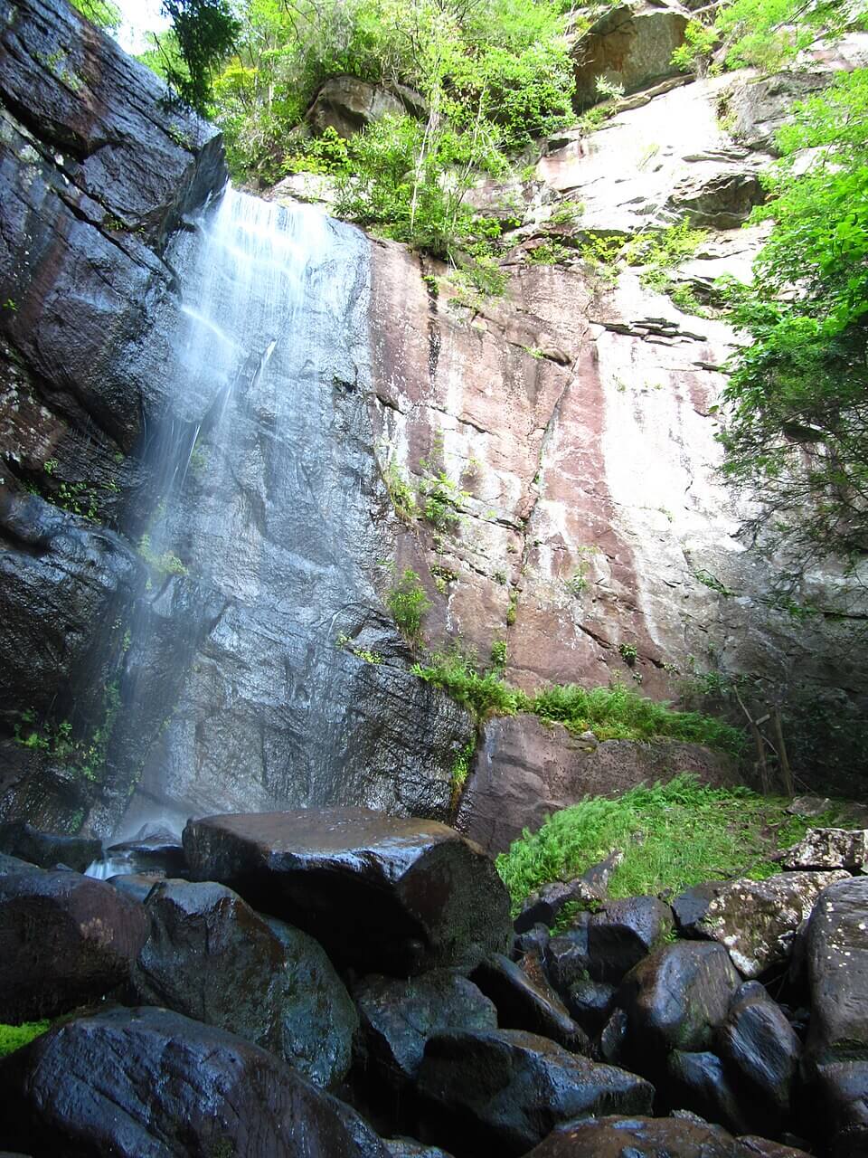 A waterfall flows down a rocky cliff surrounded by green vegetation and large dark boulders at the base, offering a scenic highlight for those enjoying Kentucky waterfall hikes.