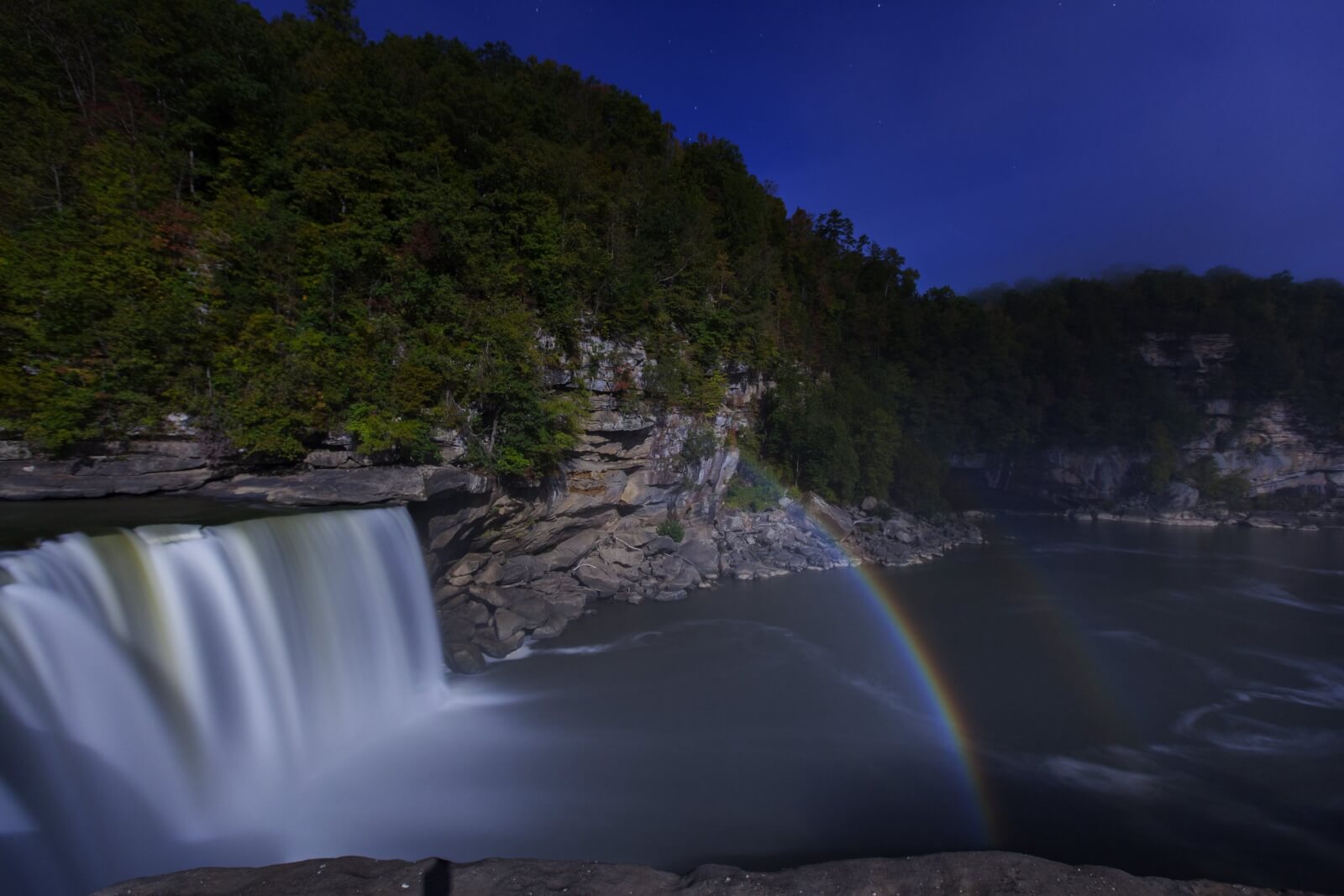 A wide waterfall cascades into a river surrounded by forested cliffs at dusk, with a faint rainbow in the mist—capturing the magic found on the best waterfall hikes during Kentucky hiking adventures.