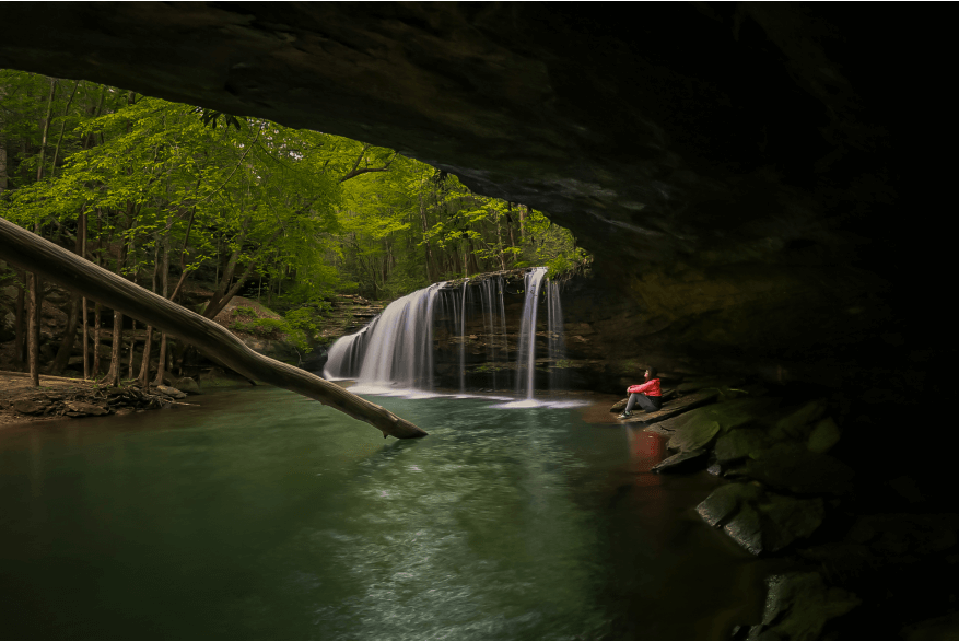 A person in a red jacket sits on rocks near a waterfall in a forest, viewed from under a rocky overhang with a fallen tree reaching toward the water—a scene typical of scenic Kentucky waterfall hikes.
