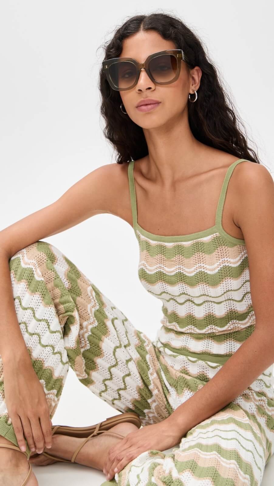 Woman wearing sunglasses, a green and white zigzag-patterned knit tank top, and matching pants, sits on the floor against a plain background—capturing effortlessly stylish summer fashion.
