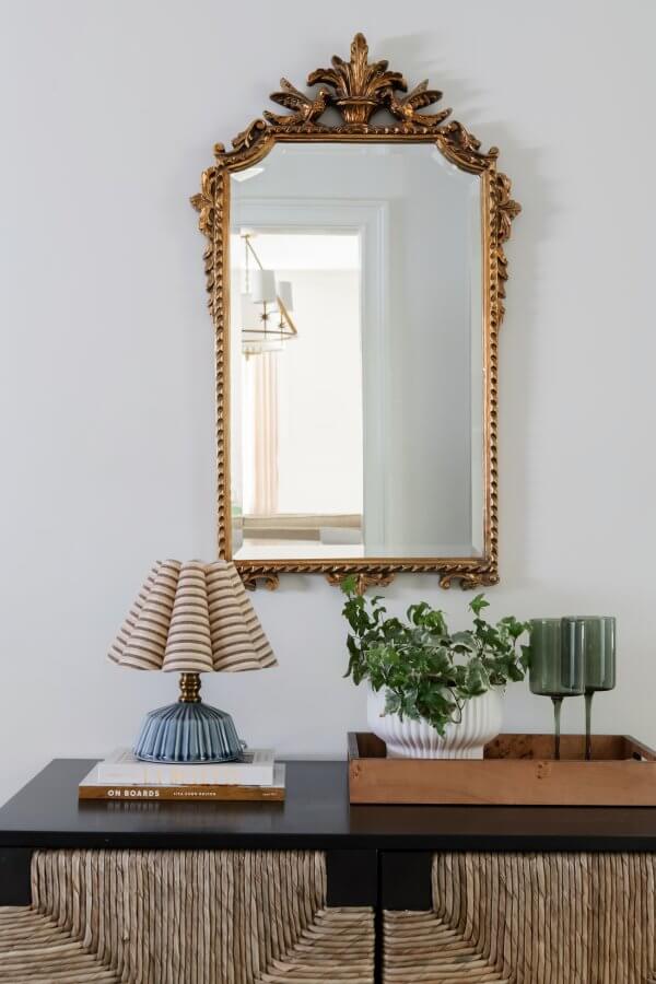 In a Sylvan Park home, an ornate gold mirror elegantly complements the woven-texture cabinet. A striped lamp, potted plant, and two green glasses on a tray complete this delightful space where traditional meets modern design.