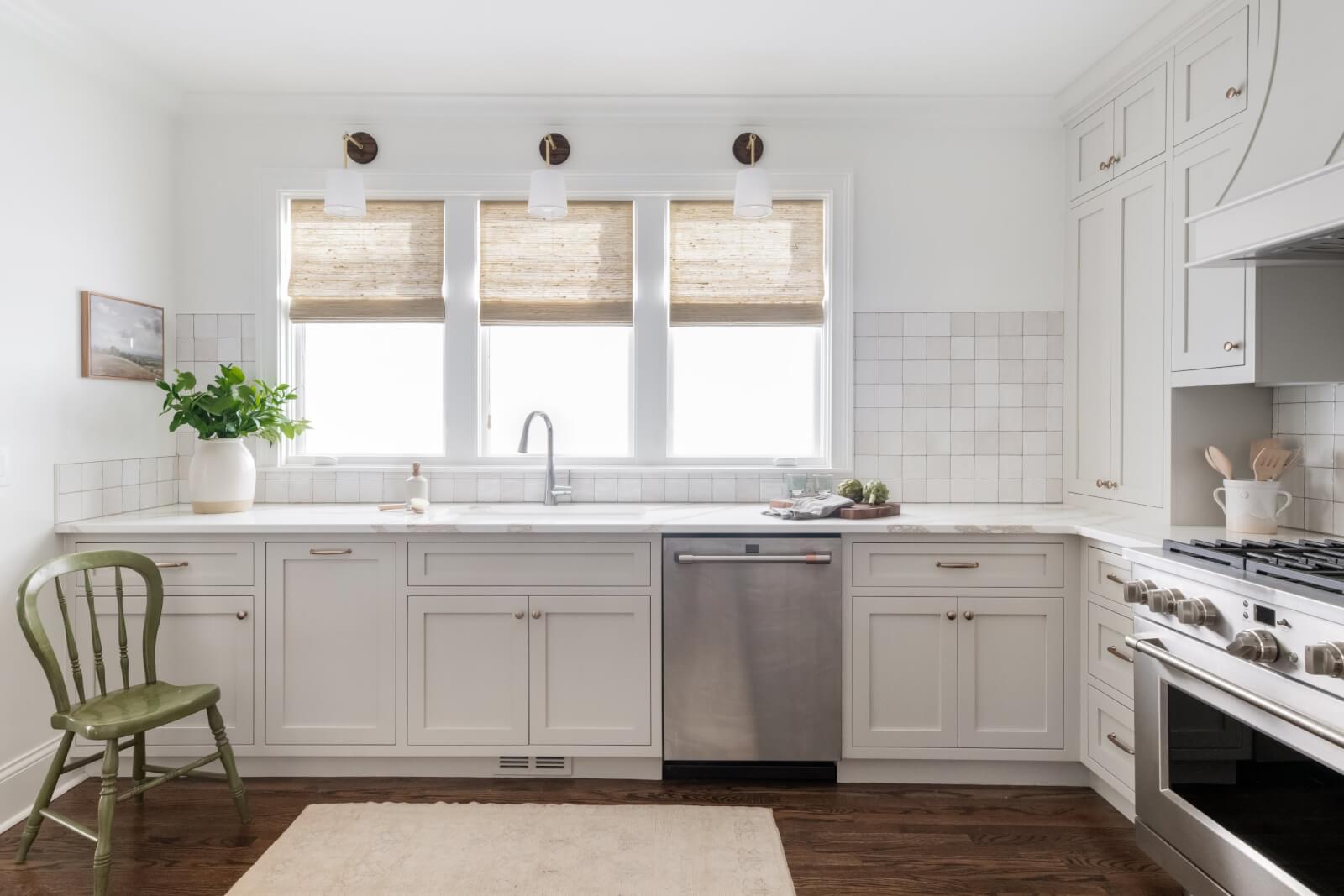 A bright Sylvan Park Home kitchen with white cabinets, a farmhouse sink, and stainless steel appliances. Three windows with woven blinds, a green chair, and a potted plant complete the modern design.