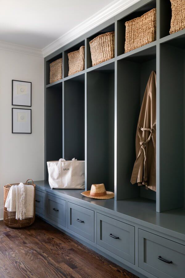 In this Sylvan Park home, a mudroom showcases modern design with dark green cabinetry, shelves adorned with wicker baskets, a coat hanging neatly alongside a tote bag and hat. Two framed pictures grace the wall while a woven basket with a blanket rests on the floor.