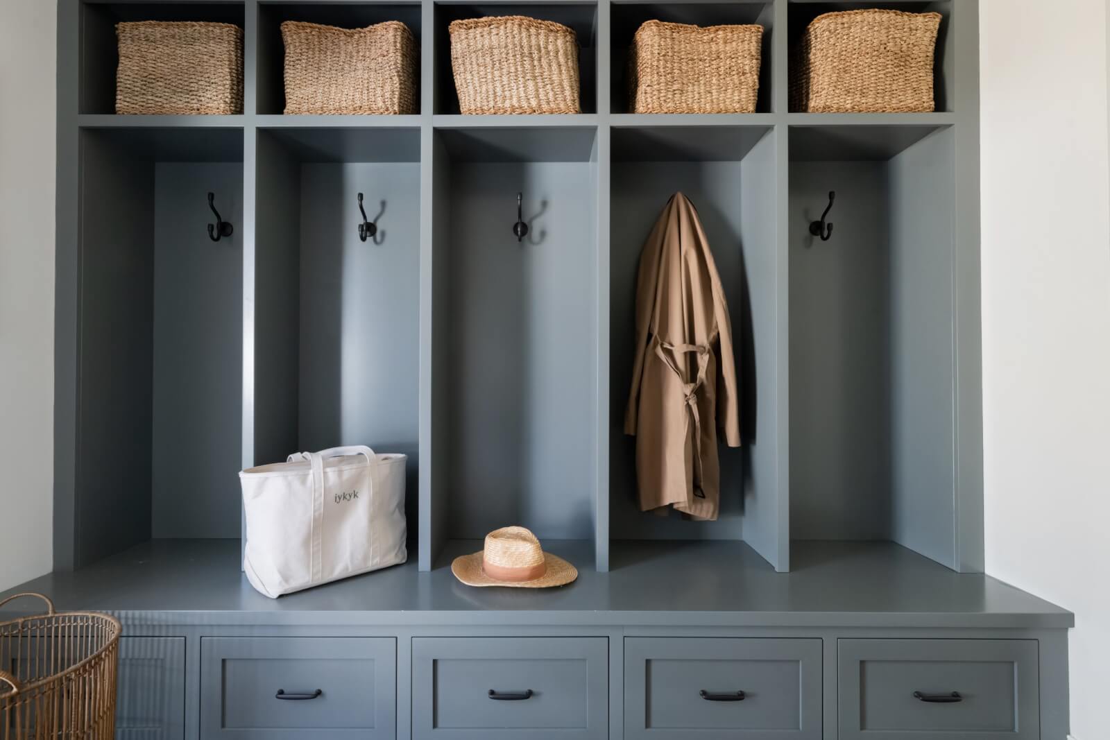 Mudroom storage unit with gray cubbies and drawers, reflecting modern design. Features three coat hooks, a hanging coat, six woven baskets, a straw hat, and a white bag on the green countertop. Perfect for adding style to your Sylvan Park home.