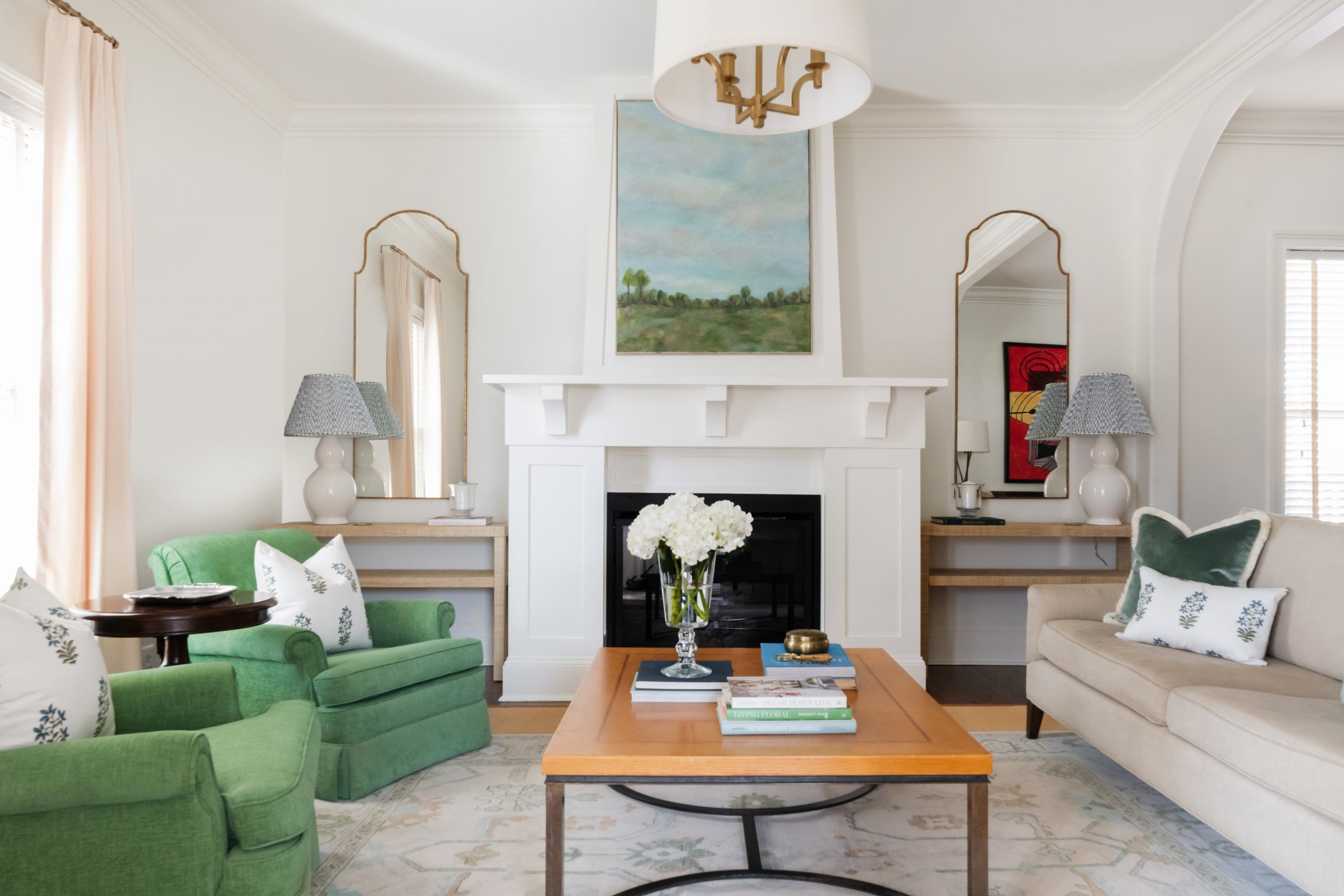 A living room in a Sylvan Park home features modern design elements, including a white fireplace, wooden coffee table, green armchairs, and beige sofa. A large painting hangs above the mantel flanked by matching lamps and mirrors.
