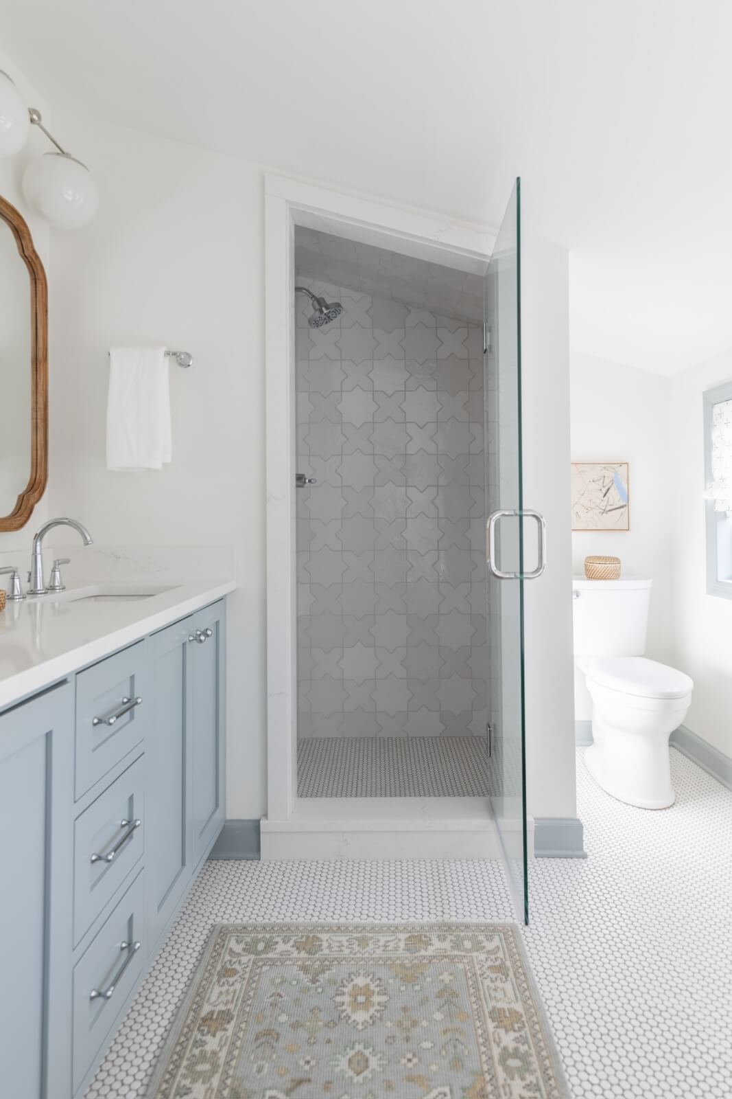 This Sylvan Park home's bathroom combines traditional meets modern design, featuring a glass shower, blue vanity with sink, and white toilet. Light-colored walls complement the decorative rug on the tiled floor, capturing the essence of Nashville charm.