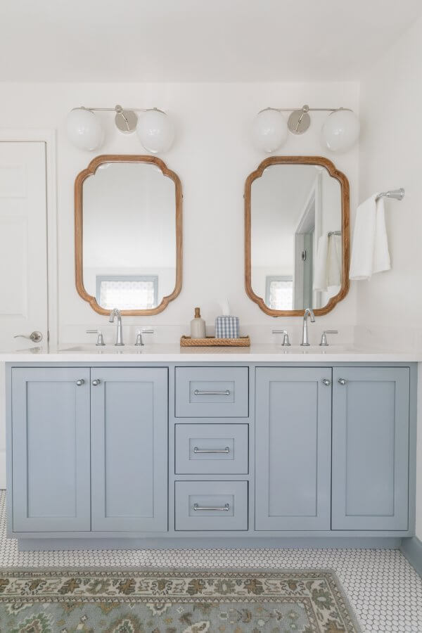 This Sylvan Park home bathroom boasts a modern design with a double vanity featuring light blue cabinets, two mirrors in wooden frames, and dual sinks. Lights illuminate above each mirror, while a cozy rug complements the patterned tile floor.