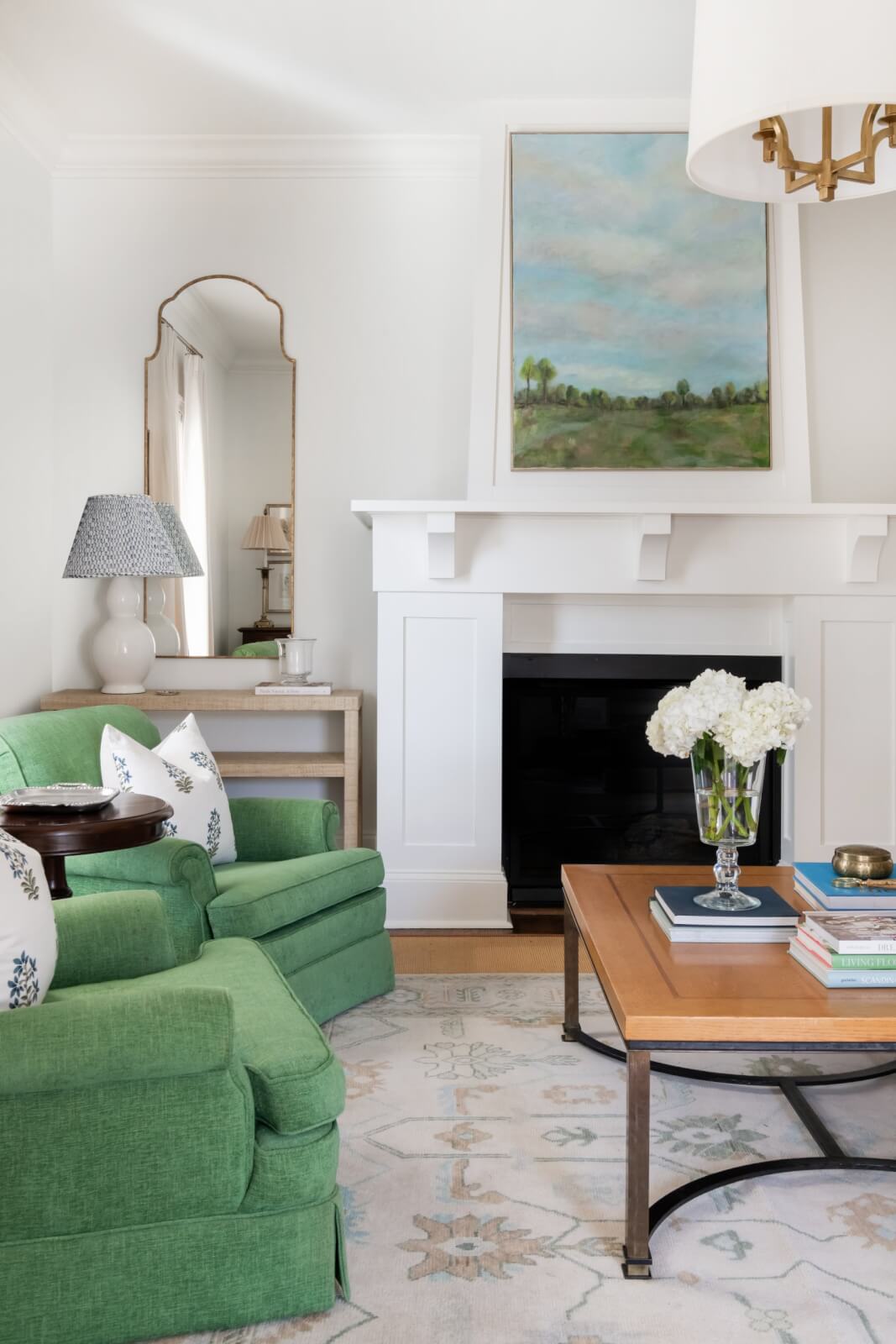 In a Sylvan Park home, the living room boasts Nashville charm with green armchairs, a wooden coffee table adorned with flowers and books, a fireplace crowned by artwork, and a mirror reflecting a lamp, seamlessly blending traditional elements with modern design.