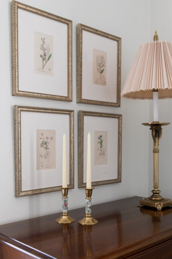 A wooden dresser with two candleholders and a lamp with a pleated shade graces this Sylvan Park home. Four framed botanical prints hang on the wall above, exuding traditional design elegance.