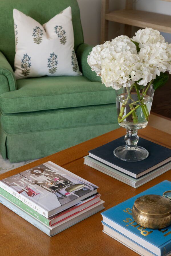 A Nashville coffee table adorned with stacked books and a gold dish is topped with a vase of white hydrangeas. In the background, a green armchair with a patterned cushion epitomizes traditional meets modern design, enhancing this Sylvan Park home's stylish harmony.