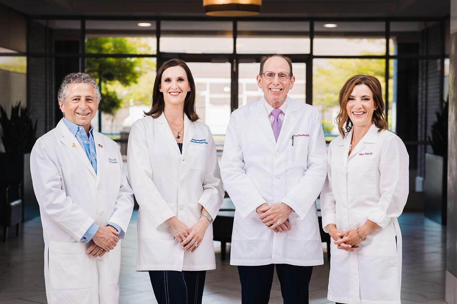 Four doctors in white lab coats, renowned as Memphis top plastic surgeons, stand together in a modern indoor setting, smiling and facing the camera.