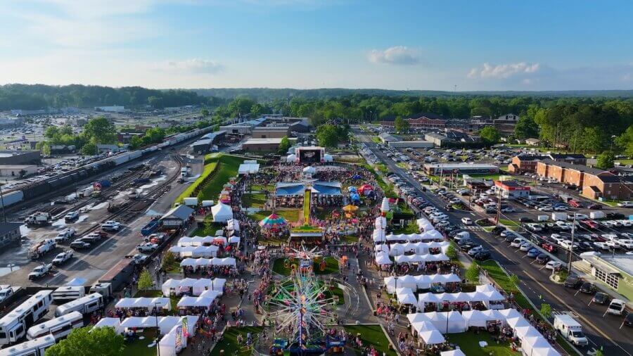 The Best Festivals in the South - 6 Aerial view of one of the best festivals in the South, featuring crowds, white tents, a Ferris wheel, amusement rides, and surrounding buildings and parking lots during the day.