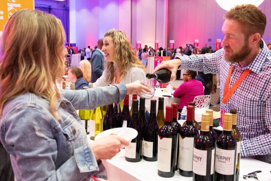 The Best Festivals in the South - 3 A man pours wine into a woman's glass at a tasting event, one of the best festivals in the South, with various wine bottles and other attendees visible in the background.