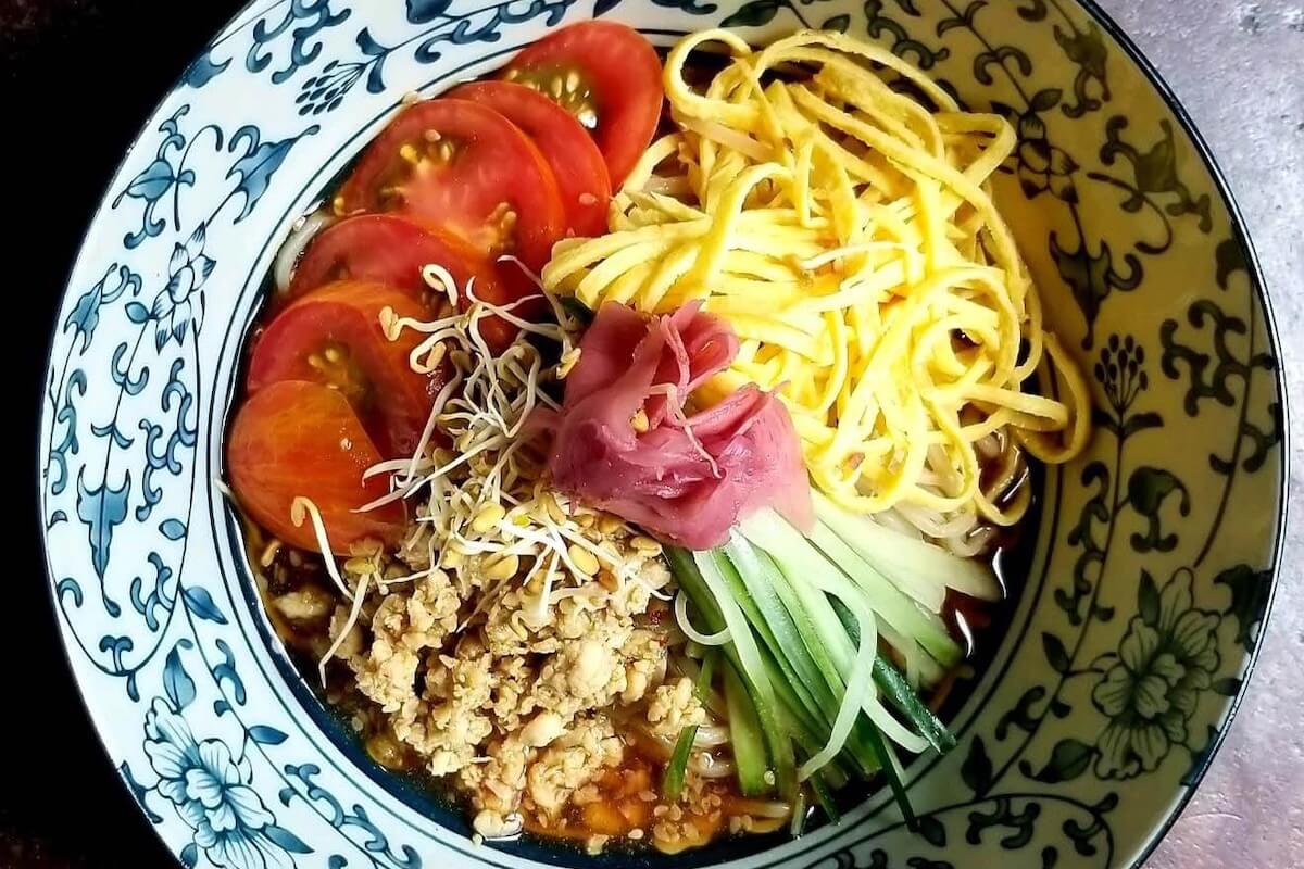 A bowl of noodle soup topped with sliced tomatoes, shredded egg, ground meat, cucumber strips, bean sprouts, and a pink pickled garnish—a delicious way to give back while dining out for life at Nashville restaurants.