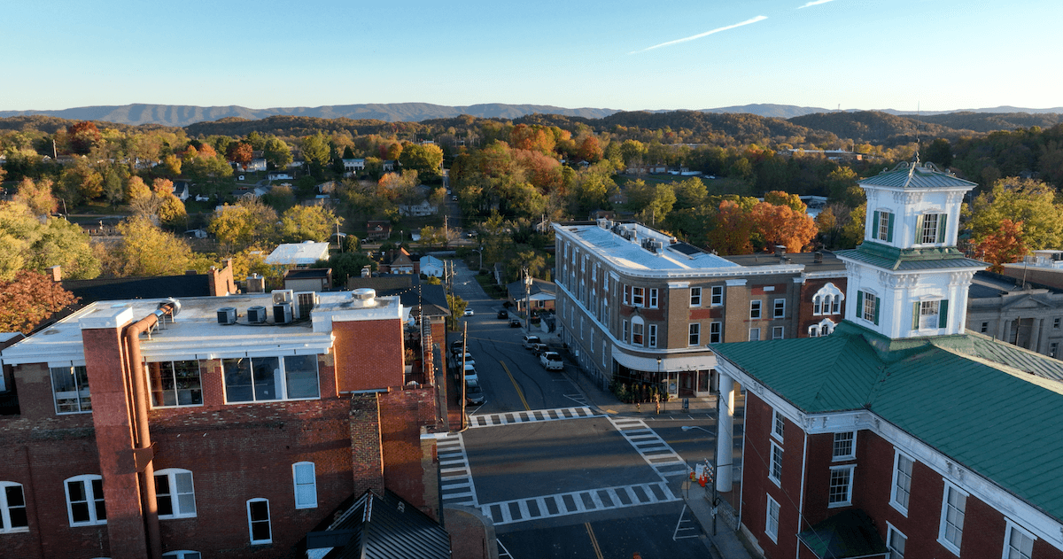 Aerial view of a small town intersection with brick buildings, tree-lined streets, and hills in the background on a clear day, capturing the charming essence of small towns in Virginia.