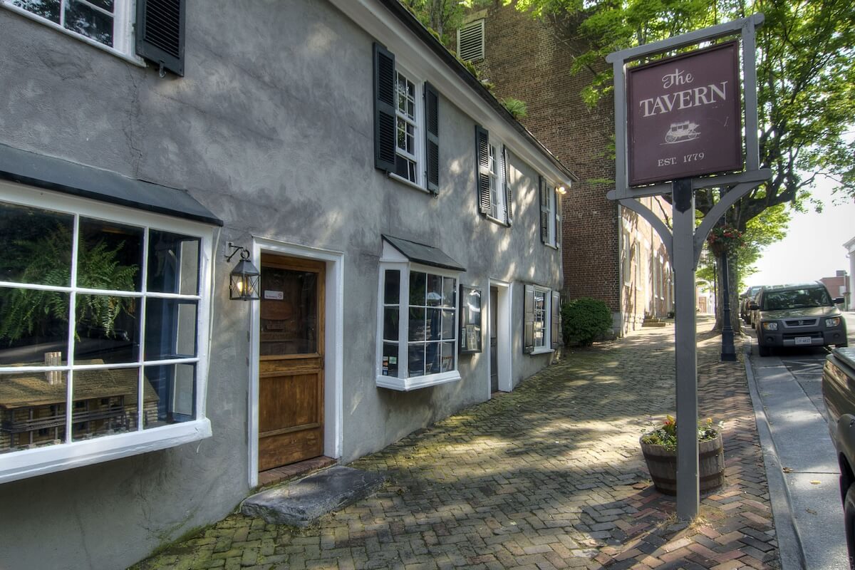 A historic stone building with white-trimmed windows and a wooden door; a sign reads "The Tavern, Est. 1779" on a brick sidewalk beside a quiet street, capturing the charm of small towns in Virginia.