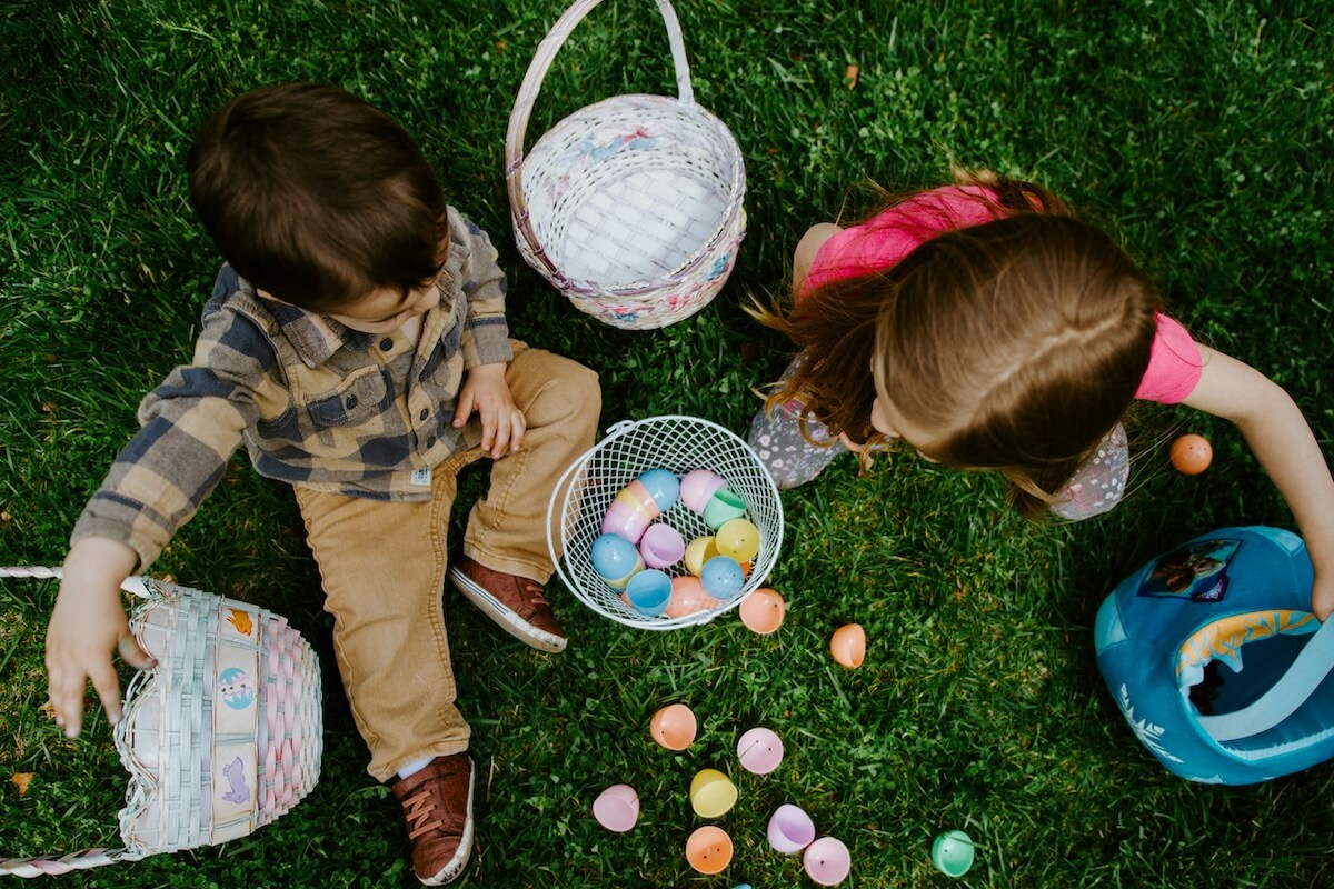 Two children sitting on grass with baskets, eagerly scanning the area during an Easter egg scavenger hunt, looking at colorful plastic eggs.