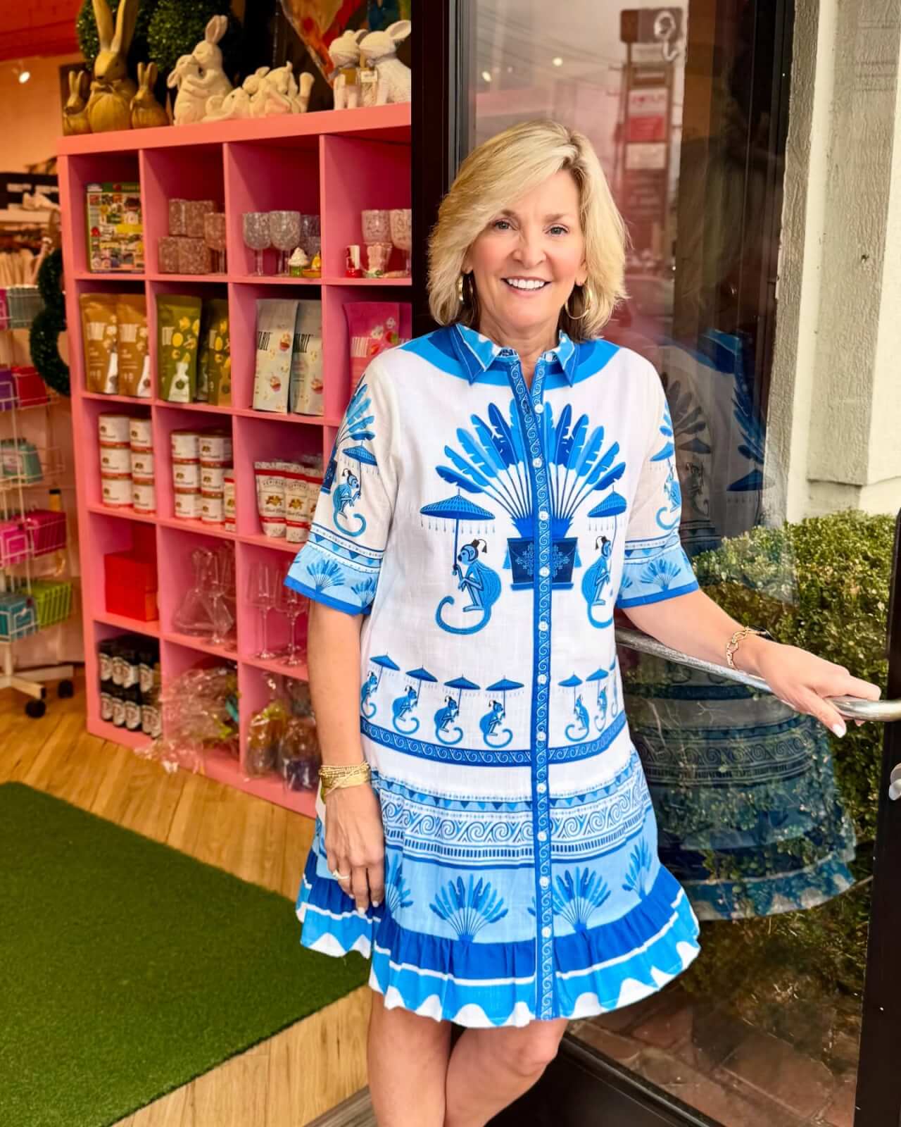 A woman in a blue and white patterned dress stands at the entrance of a shop with pink shelves, showcasing various products inside. It's one of those delightful Memphis finds that brightens up your day, reminiscent of an April afternoon spent discovering hidden gems.