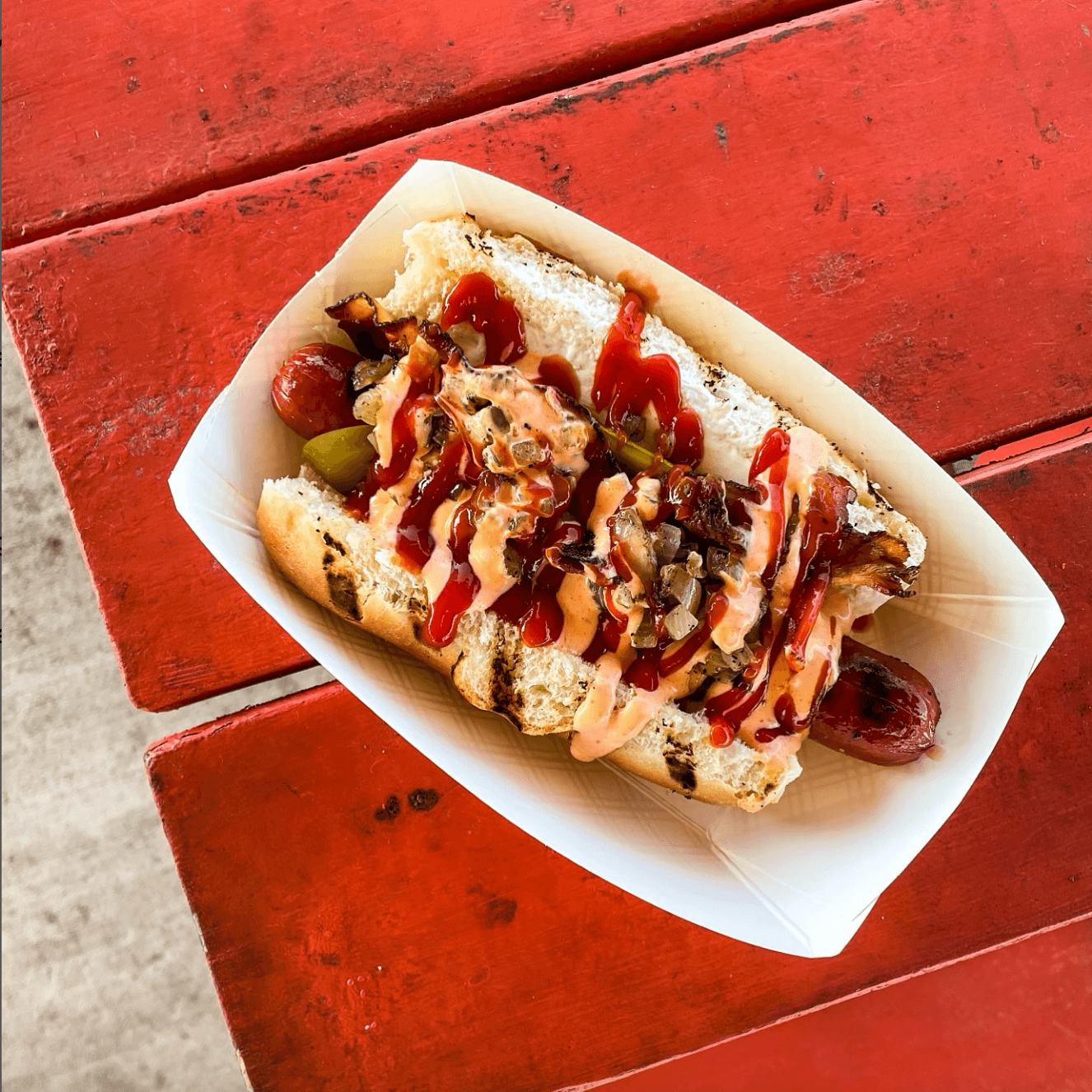 Your Updated Guide to The Nations: West Nashville's Hippest Neighborhood! - 3 On a red table sits a hot dog in a paper tray, topped with ketchup, mustard, pickles, bacon, and grilled onions.