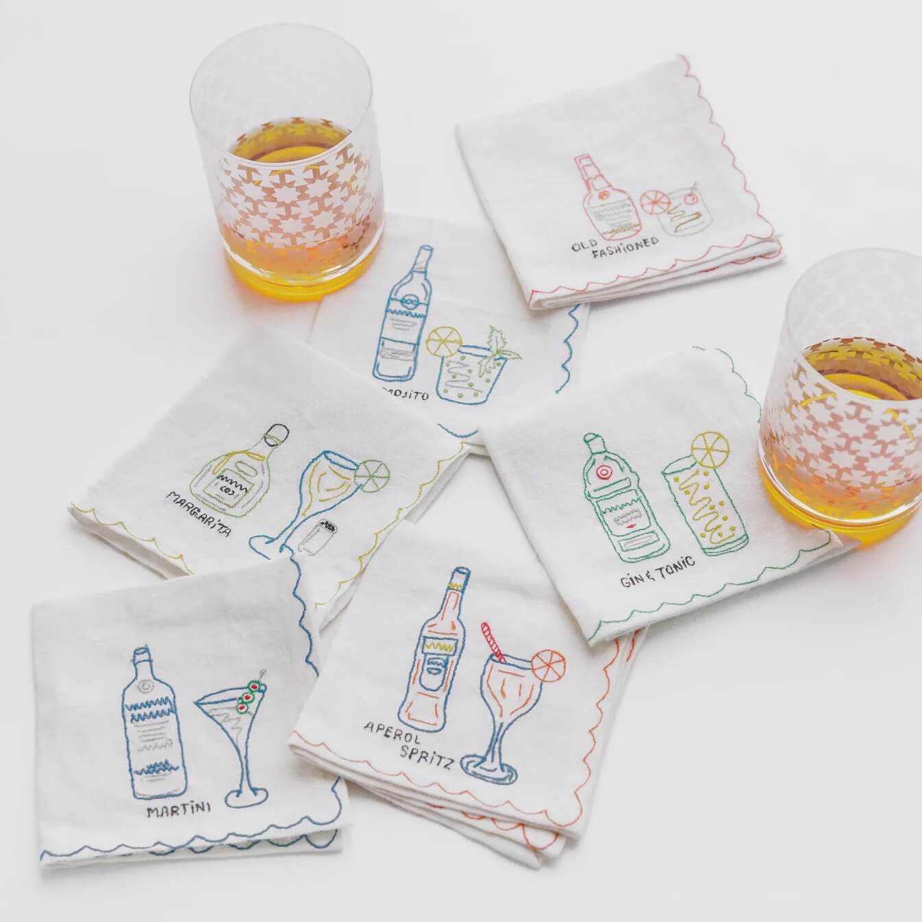 Seven embroidered cocktail-themed napkins are arranged on a white surface, with two patterned glasses containing an amber beverage placed nearby—perfect for Mother's Day gatherings or as unique Grad Gifts.