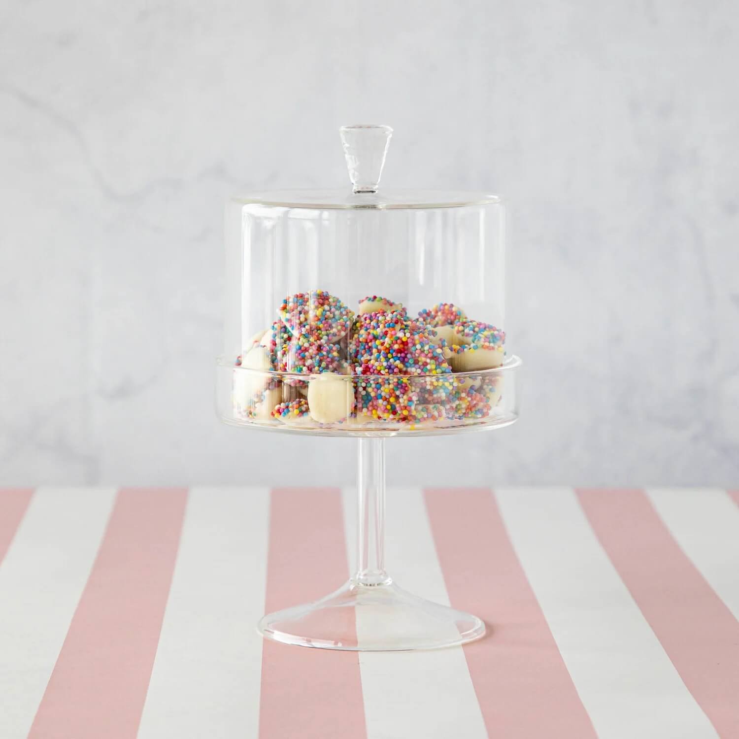 A glass cake stand with a lid displays treats covered in rainbow sprinkles, perfect for Mother's Day or Nashville grad gifts, set on a pink and white striped surface with a light gray background.