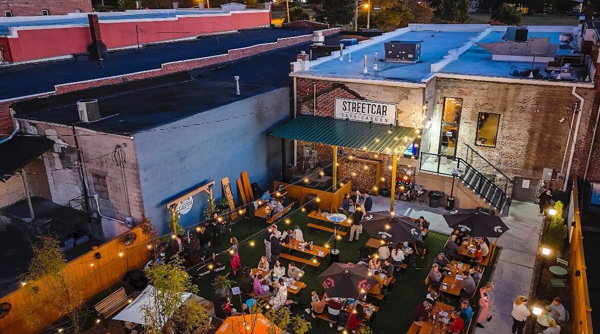 Aerial view of a vibrant outdoor dining area at Streetcar. People from various nations are seated at tables with umbrellas on a grass patio. The building combines brick and painted walls, and lights illuminate the scene.