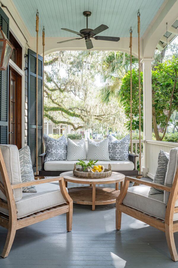 Covered porch with cushioned wooden chairs, a round coffee table with a tray of lemons, and a hanging swing sofa with patterned pillows. Ceiling fan and greenery complete this Leah Bailey design.