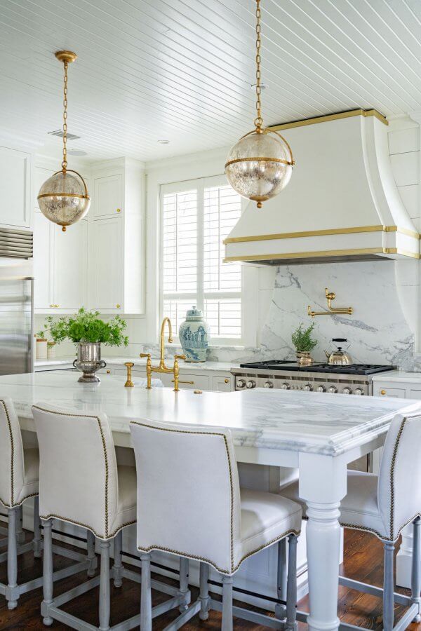 Bright, modern kitchen by Leah Bailey with white cabinetry, marble countertops, gold fixtures, upholstered barstools, and pendant lights over a large island.