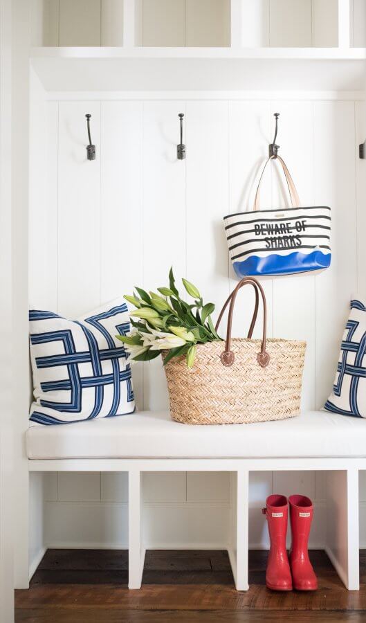 A white mudroom bench with two blue-and-white pillows, a wicker bag with flowers, a striped tote on a hook, and red rain boots on the floor create an ID Crush-worthy look inspired by Leah Bailey.