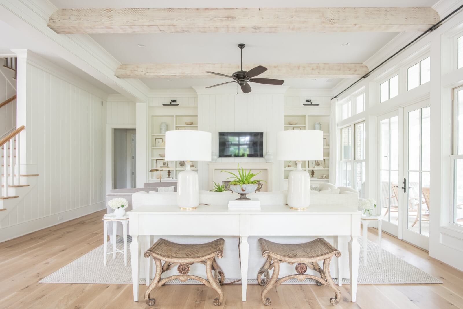 Bright living room designed by Leah Bailey, featuring white walls, wooden ceiling beams, light wood floors, neutral furniture, a TV above the fireplace, large windows, and decorative stools and lamps.