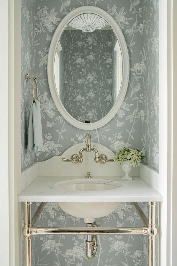 Small bathroom with floral wallpaper, oval mirror, wall-mounted faucet, white marble sink, chrome plumbing, a towel on a ring, and a small vase of flowers on the counter.