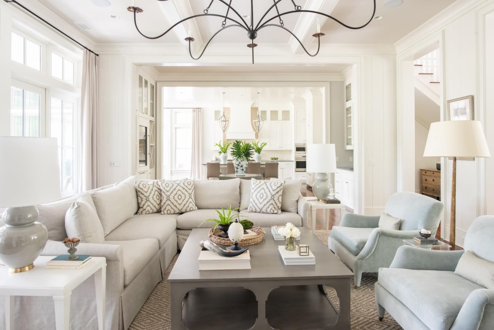 Bright living room with a beige sectional sofa, two blue armchairs, and a gray coffee table styled by Leah Bailey. Enjoy the view into a white kitchen with large windows and natural light.