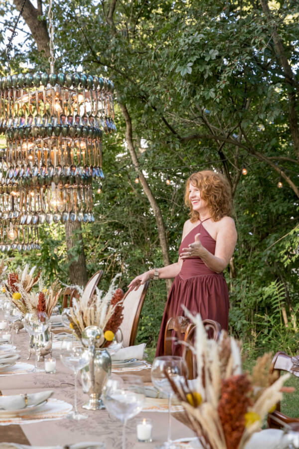 Angie Cook, in a maroon dress, stands beside a long outdoor dining table adorned with plates, glasses, and decorative arrangements beneath a unique chandelier made of spoons, her face lit by the glow of twilight.