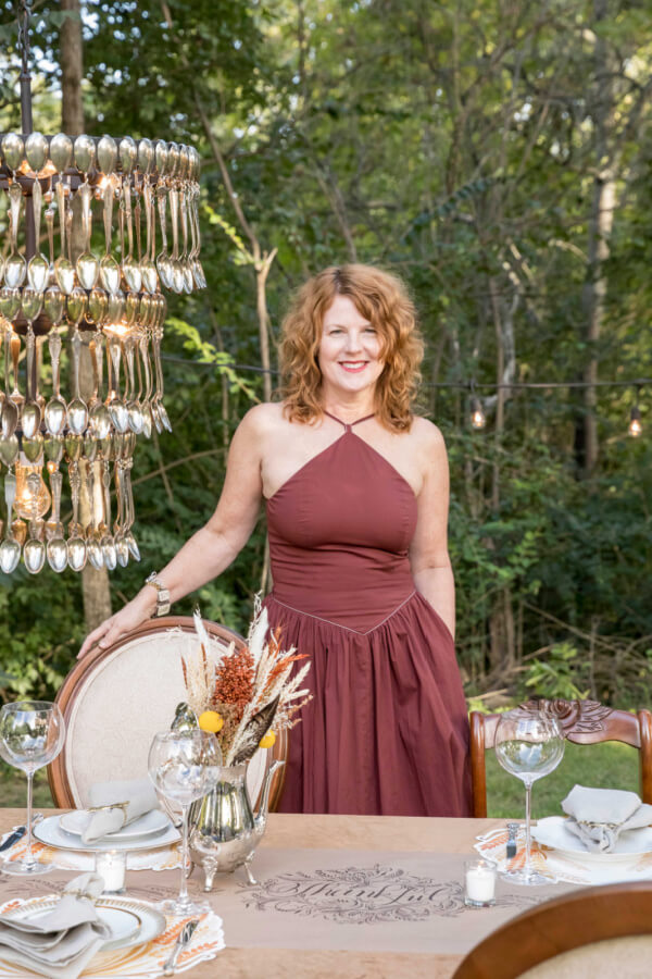 Angie Cook, in a burgundy dress, stands by an elegantly set outdoor table with a decorative spoon chandelier above, enveloped by the wooded background.
