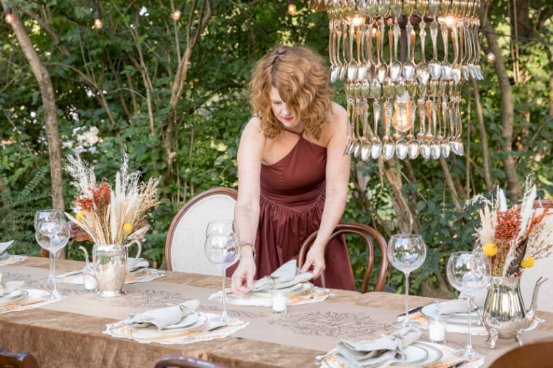Angie Cook, in a maroon dress, arranges napkins on a decorated outdoor dining table with a chandelier, surrounded by lush greenery.