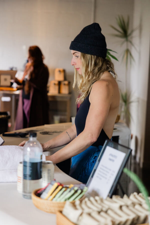 Brooke Allison, wearing a black beanie and sleeveless top, stands at a table with towels and supplies, while another person works in the background.