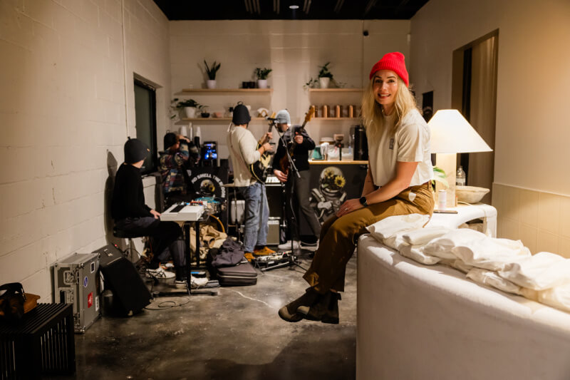 A woman in a red beanie sits on a couch, smiling at the camera as several people set up music equipment in the cozy indoor space.