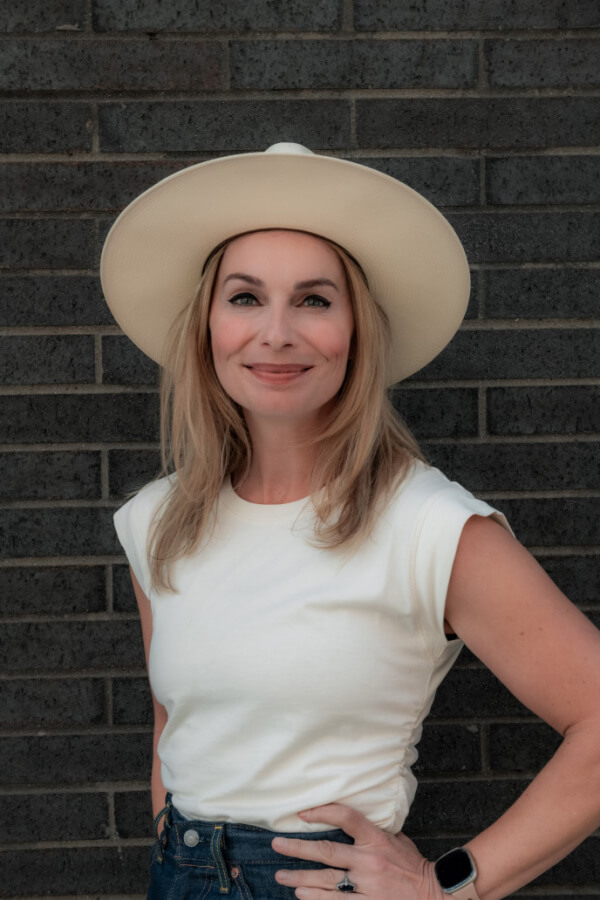 Brooke Allison, wearing a wide-brimmed hat, white sleeveless shirt, and smartwatch, stands in front of a dark brick wall, her FACE lit up with a bright smile for the camera.