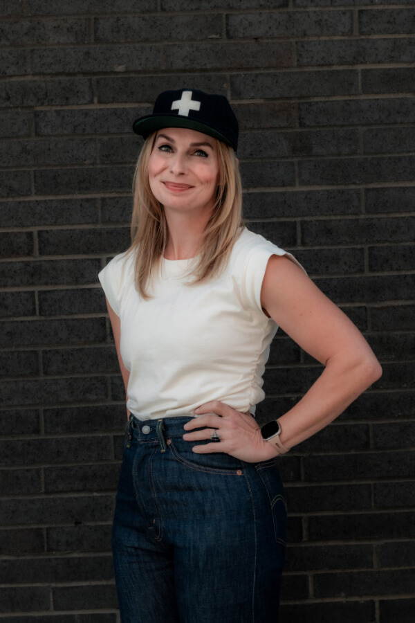 A person stands against a dark brick wall, their FACE partly shadowed, wearing a white t-shirt, blue jeans, and a black cap with a white cross.