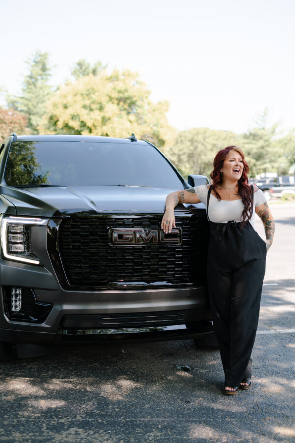 Sam McClellan of Elite Customs: Paving the Way for Women in the Auto Industry - 5 Samantha McClellan, with her striking red hair and tattoos, leans against a black GMC vehicle from Elite Customs, smiling. Trees and greenery create a serene backdrop.