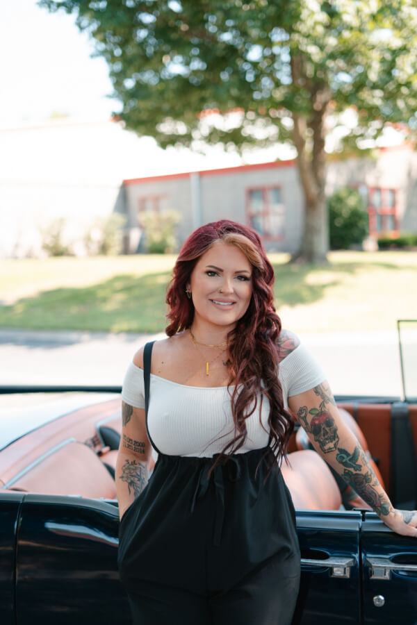 Sam McClellan of Elite Customs: Paving the Way for Women in the Auto Industry - 3 Samantha McClellan, with long red hair, stands confidently in her stylish black and white outfit before an Elite Customs convertible. In the background, a tree and building add depth to the scene.