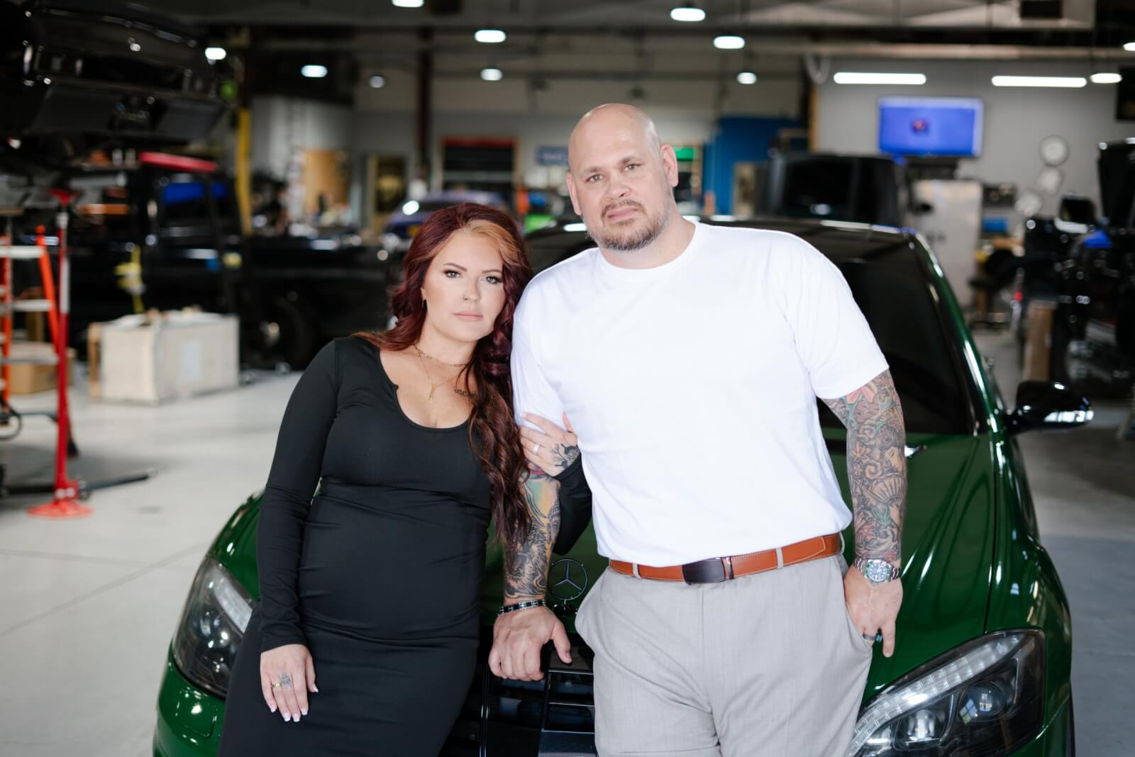 Sam McClellan of Elite Customs: Paving the Way for Women in the Auto Industry - 4 A woman and a man stand side by side, leaning against a green car in an indoor garage setting, the emblem of Elite Customs subtly gleaming on its hood. Samantha McClellan's confident stance speaks volumes as she surveys the sleek vehicle with an expert eye.