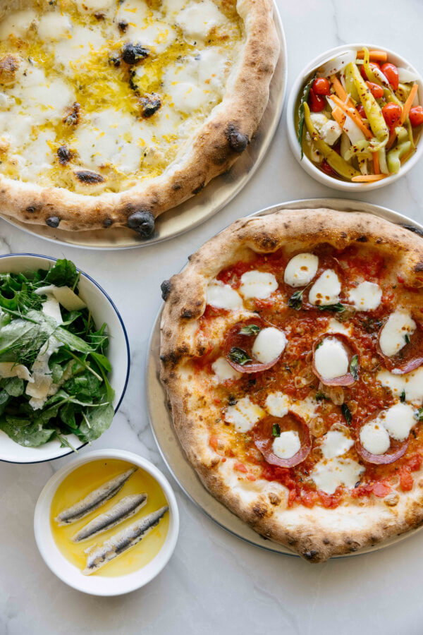 9 New Nashville Restaurants to Try This Month - 26 Two pizzas with mozzarella cheese, a bowl of mixed pickled vegetables, a green salad with parmesan, and anchovies in oil are arranged on a white surface.