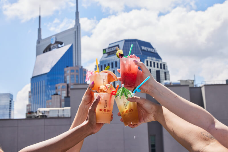 9 New Nashville Restaurants to Try This Month - 8 Four hands holding colorful cocktails with straws and garnishes are raised together in a toast, set against city buildings and a blue sky.