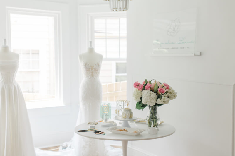 In a vibrant Nashville retail space, a bright room features two white mannequins in elegant dresses. A round table showcases flowers, stationery, and snacks. On the wall, a sign with a logo invites you to explore this exciting new retail spot.